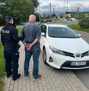 Zdjęcie przedstawia policjanta stojącego z zatrzymanym mężczyzną przed białą toyotą.