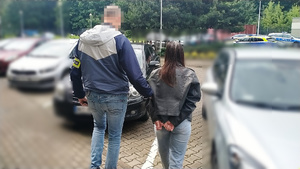 Zdjęcie przedstawia policjanta z odblaskową opaską na przedramieniu z napisem POLICJA prowadzącego po swojej prawej stronie zatrzymanego mężczyznę.