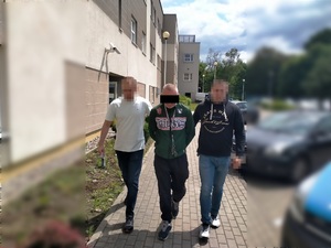 Zdjęcia przedstawia policjantów prowadzących chodnikiem pomiędzy sobą zatrzymanego ubranego w zieloną bluzę.