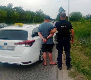 Zdjęcie przedstawia umundurowanego policjanta prowadzącego po swojej lewej stronie zatrzymanego mężczyznę.