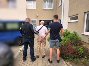 Zdjęcie przedstawia ubranych po cywilnemu policjantów, którzy prowadzą pomiędzy sobą ubranego na jasno zatrzymanego mężczyznę. Przed nimi widać okratowane okna komendy.