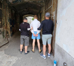 Zdjęcie przedstawia ubranych po cywilnemu policjantów, którzy prowadzą pomiędzy sobą przez przejście w podwórzu ubranego na jasno zatrzymanego mężczyznę.