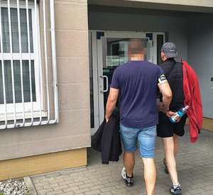 Zdjęcie przedstawia ubranego po cywilnemu policjanta, który wprowadza do budynku ubranego na ciemno zatrzymanego mężczyznę. Po ich lewej stronie widać okratowane okno komendy.