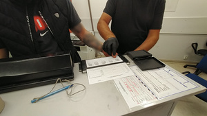 Zdjęcie przedstawia fragment sylwetki ubranego na ciemno policjanta w czarnych ochronnych rękawiczkach, który wykonuje odciski palców zatrzymanemu mężczyźnie.