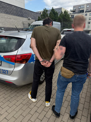 Zdjęcie przedstawia ubranego na ciemno policjanta, który prowadzi po swojej lewej stronie zatrzymanego mężczyznę. Obok nich stoi zaparkowany radiowóz.