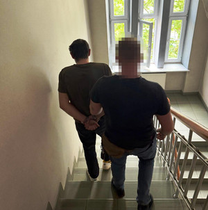 Zdjęcie przedstawia ubranego na ciemno policjanta, który sprowadza po schodach zatrzymanego mężczyznę.