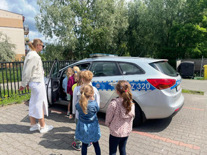 Zdjęcie przedstawia dzieci stojące w kolejce obok zaparkowanego radiowozu.
