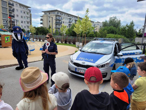 Zdjęcie przedstawia policjantkę, misia Borsuka i dzieci stojące na szkolnym boisku obok radiowozu. Policjantka trzyma w uniesionych dłoniach tarczę do zatrzymywania pojazdów.