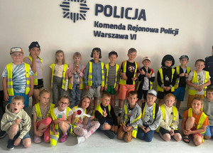 Zdjęcie przedstawia dzieci ubrane w odblaskowe kamizelki i stojące pod białą ścianą z granatowym napisem KOMENDA REJONOWA POLICJI WARSZAWA VI