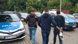 Zdjęcie przedstawia policjantów prowadzących przez parking zatrzymanego mężczyznę.