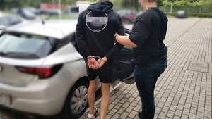Zdjęcie przedstawia policjanta prowadzącego do samochodu zatrzymanego mężczyznę. Mężczyzna ten ma kajdanki założone na rękach trzymanych z tyłu.