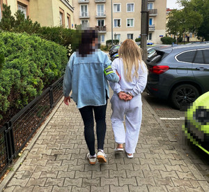 Zdjęcie przedstawia policjantkę ubraną niebieską kurtkę jeansową, która prowadzi po chodniku zatrzymaną kobietę. Kobieta ta ma kajdanki założone na rękach trzymanych z tyłu.