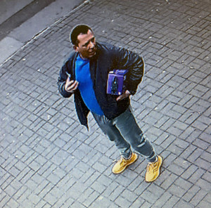 Zdjęcie przedstawia mężczyznę stojącego na chodniku i trzymającego w lewej ręce kwadratowy pojemnik. Mężczyzna jest ubrany w ciemną kurtkę, pod nią ma niebieską bluzkę, niebieskie spodnie jeansowe i żółte buty sportowe.