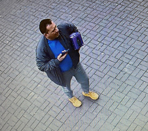 Zdjęcie przedstawia mężczyznę stojącego na chodniku i trzymającego w lewej ręce kwadratowy pojemnik. Mężczyzna jest ubrany w ciemną kurtkę, pod nią ma niebieską bluzkę, niebieskie spodnie jeansowe i żółte buty sportowe.
