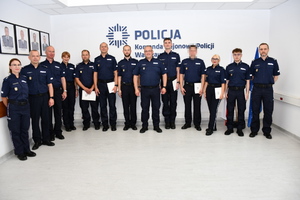 Zdjęcie przedstawia policjantów stojących w jednym rzędzie pod ścianą z napisem KOMENDA REJONOWA POLICJI WARSZAWA VI.