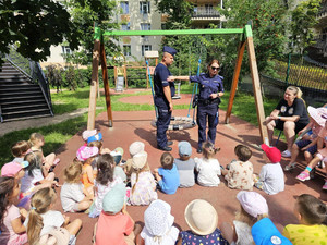 Zdjęcie przedstawia policjantów stojących przy radiowozie. Przed nimi na ławkach siedzą dzieci.