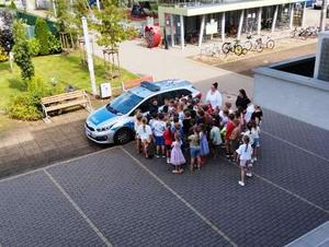 Zdjęcie przedstawia policjantkę i dzieci stojące przy zaparkowanym radiowozie.