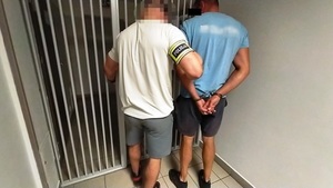 Zdjęcie przedstawia ubranego na jasno policjanta, z opaską POLICJA na prawym przedramieniu, który wyprowadza z pomieszczenia dla osób zatrzymanych mężczyznę ubranego w niebieską koszulkę.
