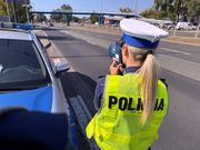 Zdjęcie przedstawia policjantkę z Wydziału Ruchu Drogowego stojącą przy zaparkowanym radiowozie i kontrolującą prędkość pojazdów.