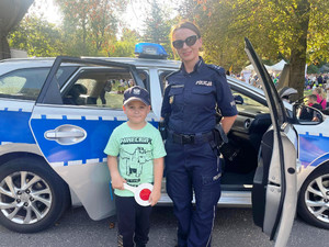 Zdjęcie przedstawia policjantkę stojącą przy dziecku. Za nimi stoi zaparkowany radiowóz.