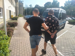 Zdjęcie przedstawia ubranego w ciemną koszulkę i niebieskie spodenki jeansowe policjanta, który prowadzi po dziedzińcu zatrzymanego mężczyznę. Mężczyzna ten ma kajdanki założone na rękach trzymanych z tyłu.