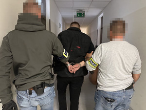 Zdjęcie przedstawia dwóch policjantów ubranych w cywilne ubrania, którzy prowadzą pomiędzy sobą zatrzymanego mężczyznę.