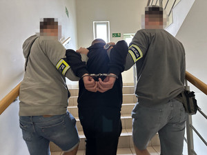 Zdjęcie przedstawia dwóch policjantów ubranych w cywilne ubrania, którzy prowadzą pomiędzy sobą zatrzymanego mężczyznę.