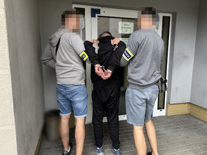 Zdjęcie przedstawia dwóch policjantów ubranych w cywilne ubrania, którzy prowadzą pomiędzy sobą zatrzymanego mężczyznę.