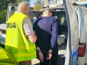 Zdjęcie przedstawia policjanta ubranego w odblaskową kamizelkę z napisem POLICJA, który umieszcza w radiowozie zatrzymanego mężczyznę.