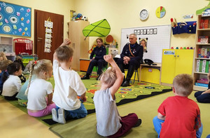Zdjęcie przedstawia policjantkę i mężczyznę w mundurze siedzących przed dziećmi siedzącymi na dywanie.