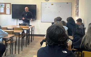 Zdjęcie przedstawia mężczyznę stojącego w klasie przed uczniami siedzącymi w ławkach.