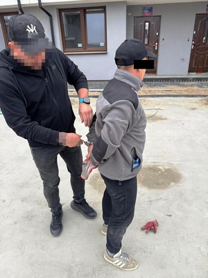 Zdjęcie przedstawia policjanta w czarnej czapce z daszkiem, który stoi za zatrzymanym mężczyzną i zakłada mu kajdanki na ręce trzymane z tyłu.