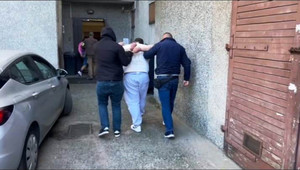 Zdjęcie przedstawia policjantów ubranych na ciemno, którzy prowadzą po dziedzińcu, pomiędzy sobą zatrzymanego mężczyznę.