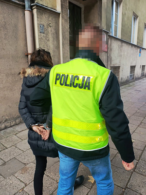 Zdjęcie przedstawia policjanta ubranego w odblaskową kamizelkę, który prowadzi po chodniku, wzdłuż budynku ubraną na ciemno, zatrzymaną kobietę. Ma ona kajdanki założone na rękach trzymanych z tyłu.
