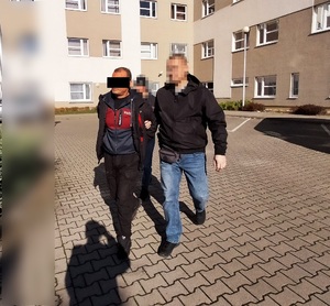 Zdjęcie przestawia ubranego na ciemno policjanta, z komputerowym filtrem zasłaniającym jego twarz, który prowadzi przez dziedziniec, po swojej prawej stronie zatrzymanego mężczyznę. Cześć twarzy mężczyzny zasłania czarny prostokąt.