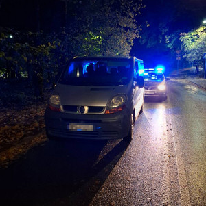 Zdjęcie jest wykonane nocą. Widać zaparkowany pojazd m-ki Renault i stojący za nim z zapalonymi sygnałami świetlnymi radiowóz.