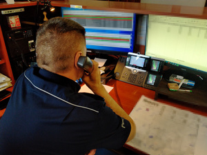 Zdjęcie przedstawia umundurowanego policjanta siedzącego przed dwoma monitorami komputera i trzymającego słuchawkę telefonu przy prawym uchu.