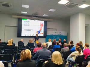 Zdjęcie przedstawia kobietę stojącą przed siedzącymi na krzesłach ludźmi.