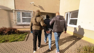 Zdjęcie przedstawia dwóch policjantów, którzy prowadzą pomiędzy sobą, po chodniku zatrzymanego mężczyznę.