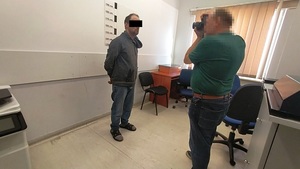 Zdjęcie przedstawia policjanta, który wykonuje zdjęcia zatrzymanemu mężczyźnie stojącemu pod ścianą z czarną skalą.