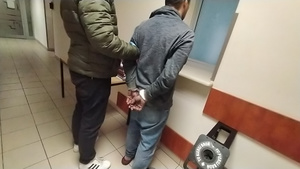 Zdjęcie przedstawia policjanta, który stoi z zatrzymanym mężczyzną przed zamkniętym okienkiem do pomieszczenia oficera dyżurnego.