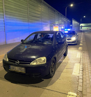 Zdjęcie przedstawia ciemnego opla stojącego na pasie ruchu, za którym stoi radiowóz z włączonymi sygnałami świetlnymi.