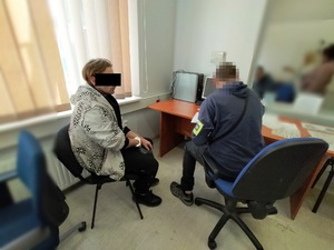 Zdjęcie przedstawia policjanta siedzącego przy biurku, który przesłuchuje mężczyznę. Zatrzymany mężczyzna ma kajdanki na rekach trzymanych z tyłu, a część jego twarzy na wysokości oczu zasłania czarny prostokąt.