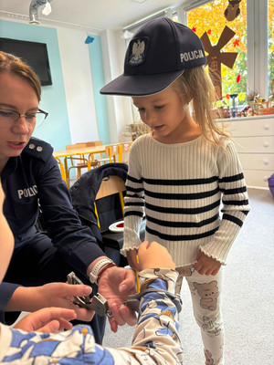 Zdjęcie przedstawia policjantkę stojącą z dziećmi. Jedno dziecko ma głowie policyjną czapkę z daszkiem i trzyma w dłoniach kajdanki.