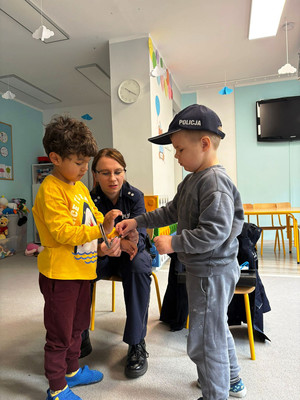 Zdjęcie przedstawia policjantkę z dziećmi. Jedno dziecko ma na głowie policyjną czapkę z daszkiem i trzyma w dłoniach, razem z drugim dzieckiem kajdanki.