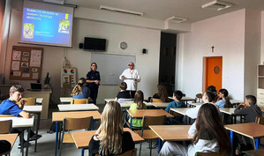 Zdjęcie przedstawia policjantkę i ubranego na jasno mężczyznę, którzy stoją na przodzie klasy, przed siedzącymi w ławkach uczniami. Za nimi, na ścianie widać kolorowy slajd prezentacji.
