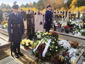Zdjęcie przedstawia dwóch policjantów w umundurowaniu wyjściowym stojących po obu stronach pomnika na cmentarzu.