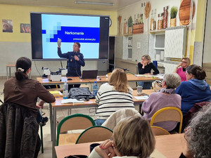 Zdjęcie przedstawia policjantkę stojącą przed ekranem z prezentacją i trzymającą w uniesionej dłoni małą torebeczkę.