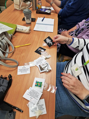 Zdjęcie przedstawia sylwetki dwóch osób siedzących w ławce, na której leżą różne przedmioty umieszczone w małych torebeczkach.