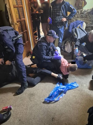 Zdjęcie przedstawia policjanta pochylającego się nad mężczyzną leżącym na dywanie. W tle widać innych policjantów w ubraniach cywilnych.